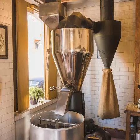 A large coffee grinder inside a building with a burlap coffee bag hanging in the background.