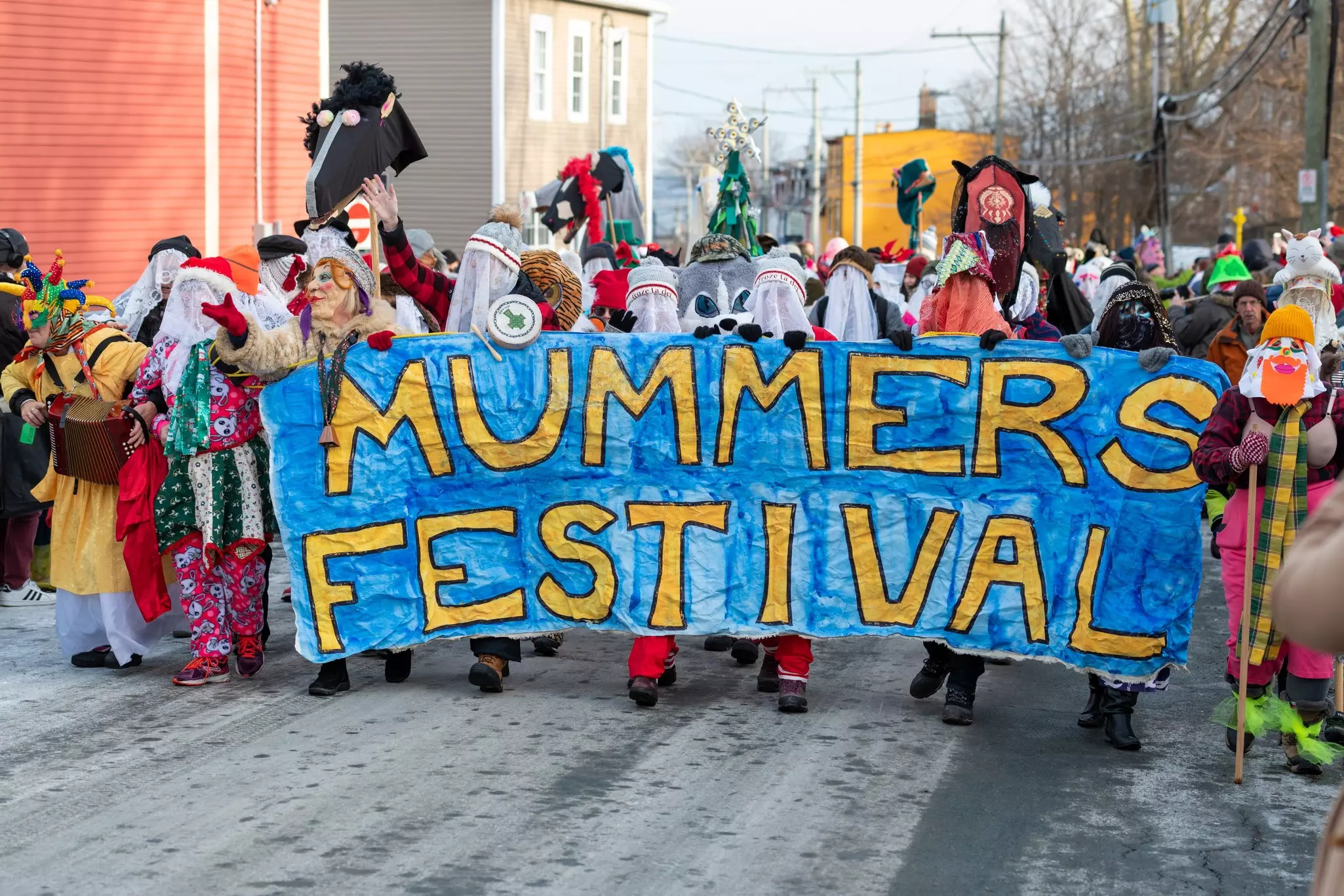 People in a parade dressed in colorful costumes and face coverings, carrying a large, handmade, blue sign reading "Mummers Festival" in blue hand-painted letters.