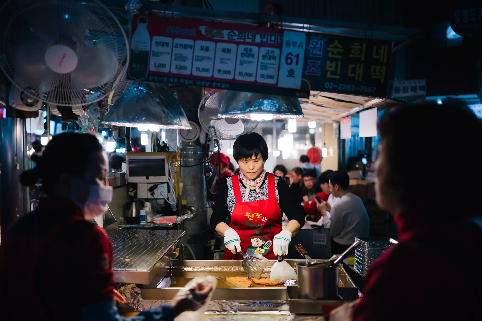 A food vendor prepares meat on a hot plate in a busy food market.