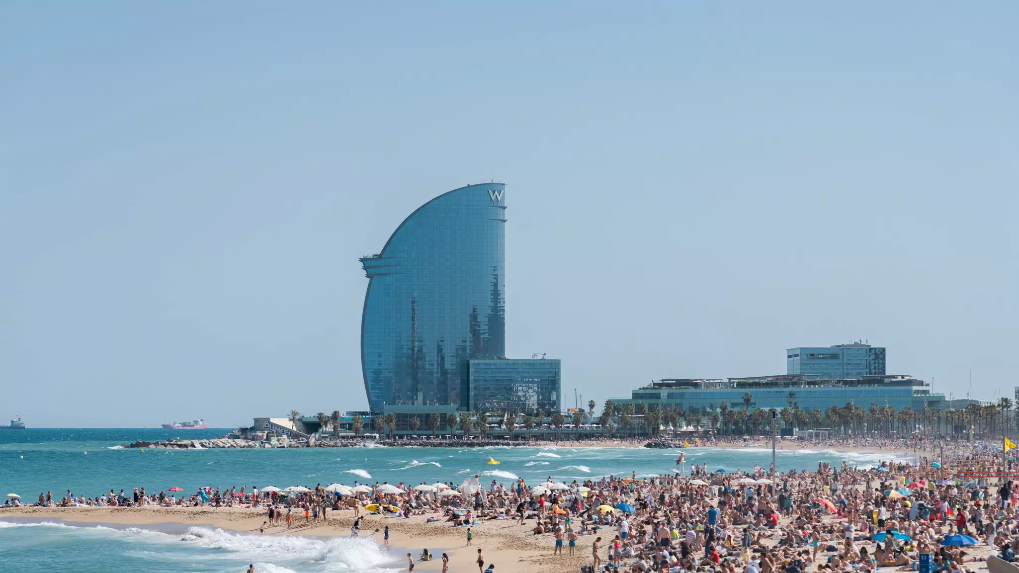 A busy beach with a fin-shaped glass hotel in the distance.