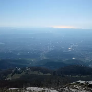 View from a rocky mountain summit over dark forested ridges to a wide hazy plain filled with a sprawling city and a reservoir.