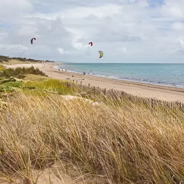 A grass-covered sand dune at the edge of a beach where people are kite surfing.