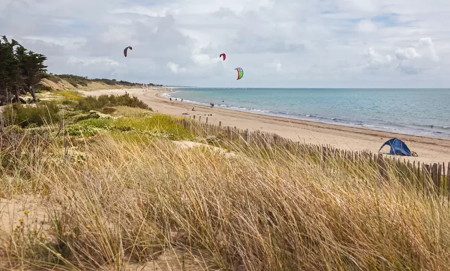 A grass-covered sand dune at the edge of a beach where people are kite surfing.