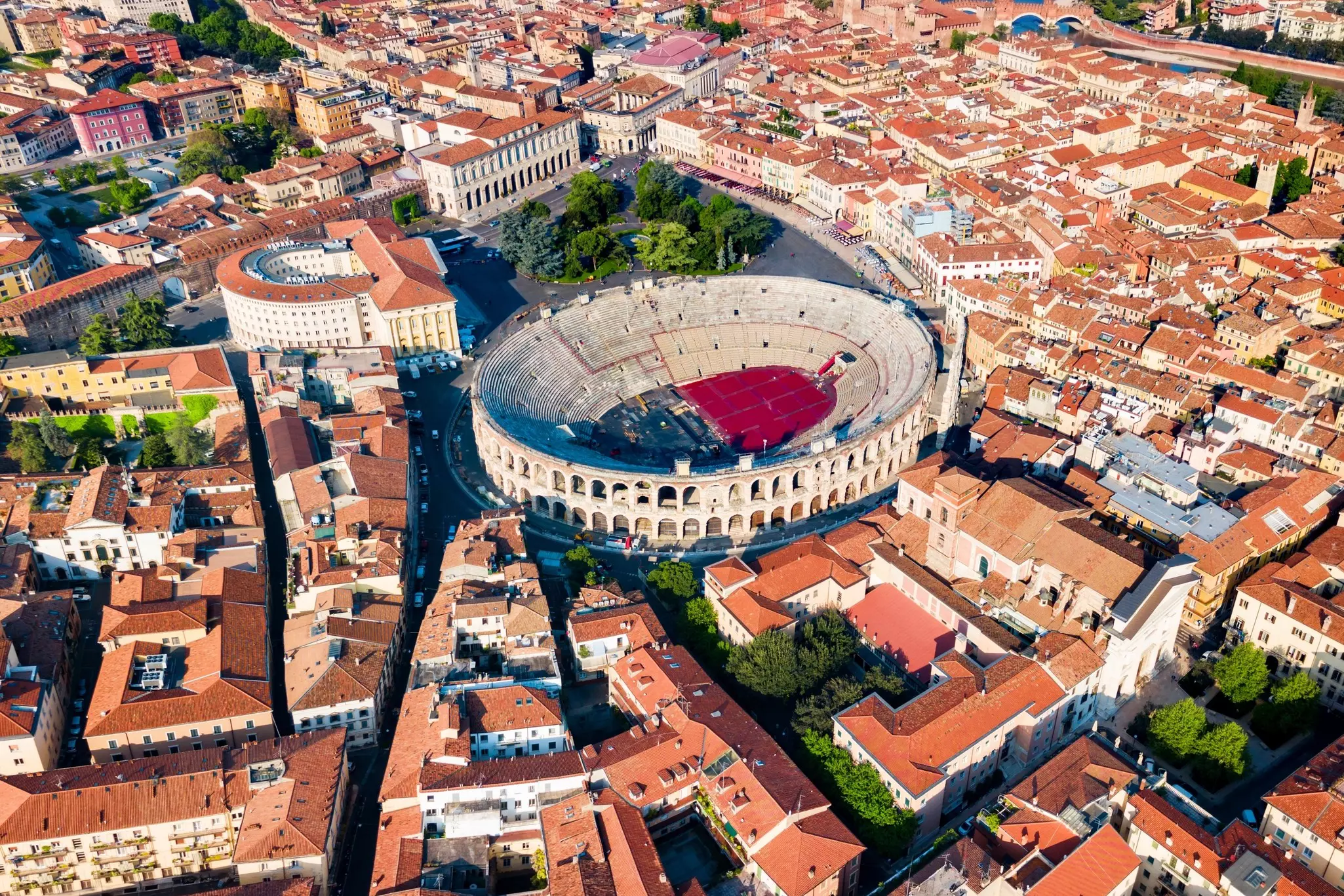 An aerial view of a large round amphitheater in the middle of a historic city, with buildings showing red-tiled roofs.