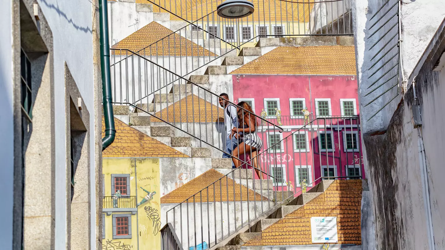 A couple walk up a set of stairs covered in street art depicting the sides of buildings and rooftops, creating an effect where it seems as though the couple is walking on roofs