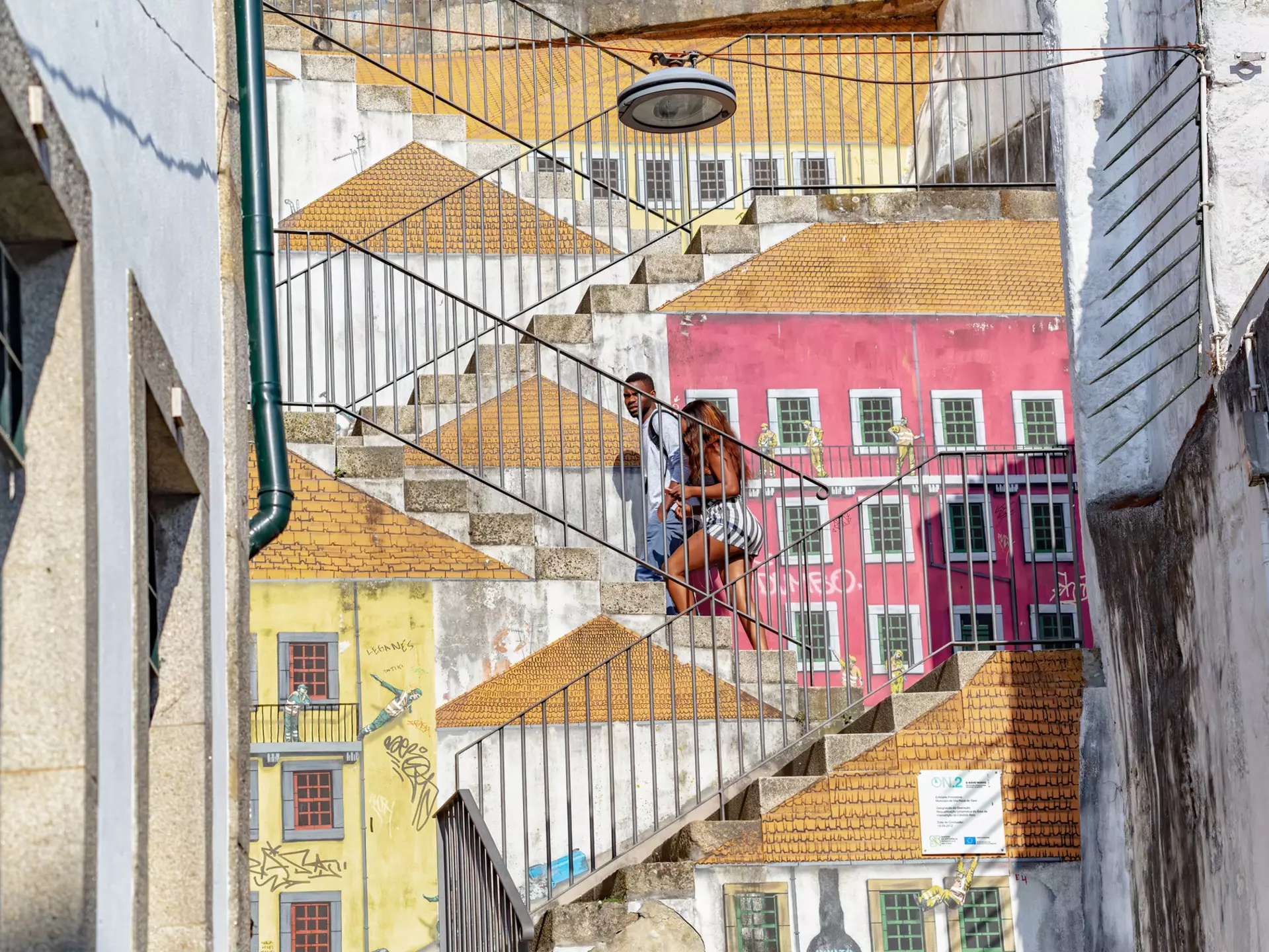 A couple walk up a set of stairs covered in street art depicting the sides of buildings and rooftops, creating an effect where it seems as though the couple is walking on roofs
