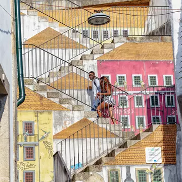 A couple walk up a set of stairs covered in street art depicting the sides of buildings and rooftops, creating an effect where it seems as though the couple is walking on roofs