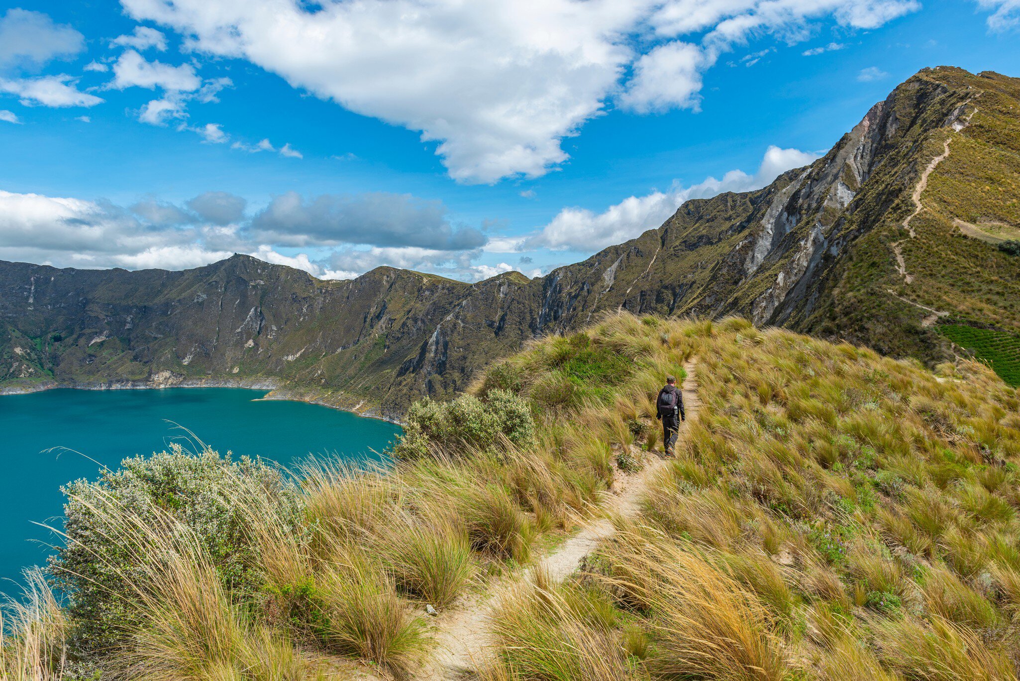 A person with a backpack walks a narrow path through tall grass beside a turquoise lake ringed by steep slopes