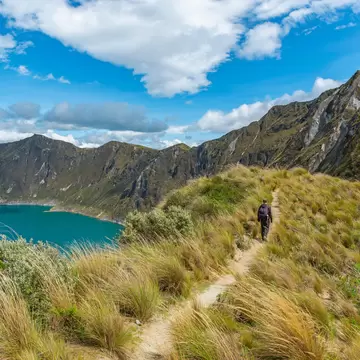 A person with a backpack walks a narrow path through tall grass beside a turquoise lake ringed by steep slopes
