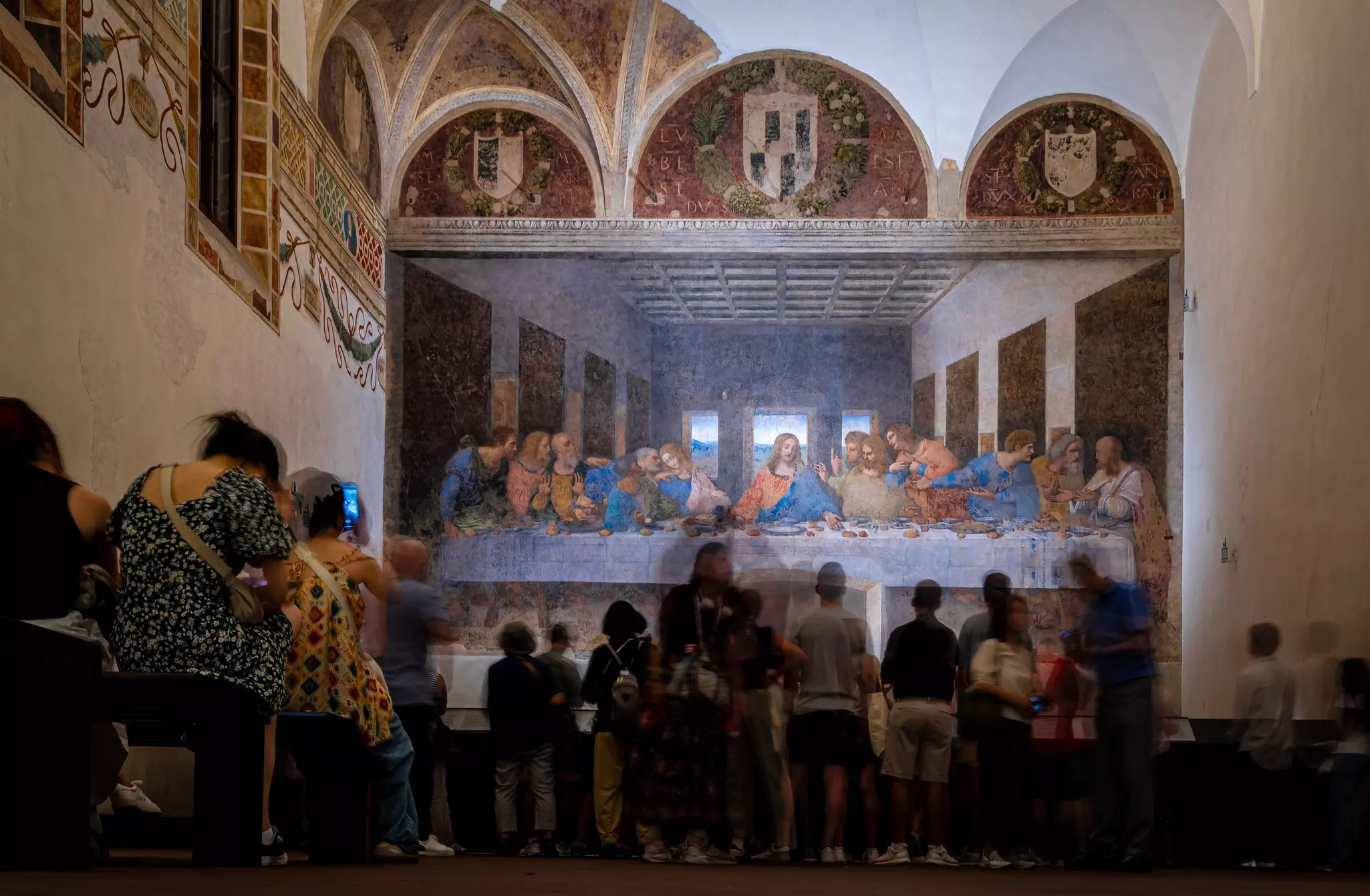 People are seen from behind admiring a fresco covering the wall of a church building.