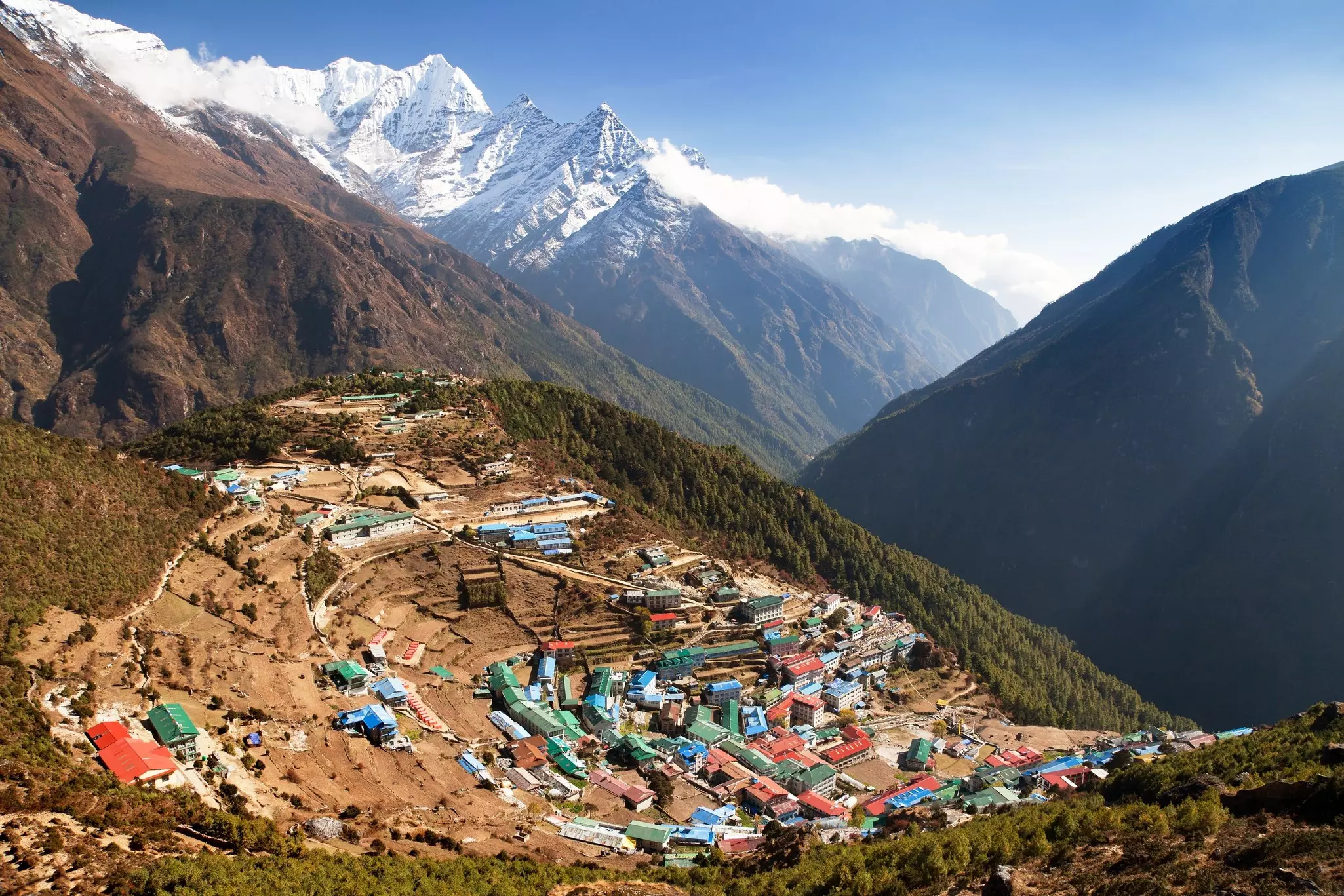 A mountain village surrounded by woodland, with small huts with red, blue and green roofs tucked into the hillside.