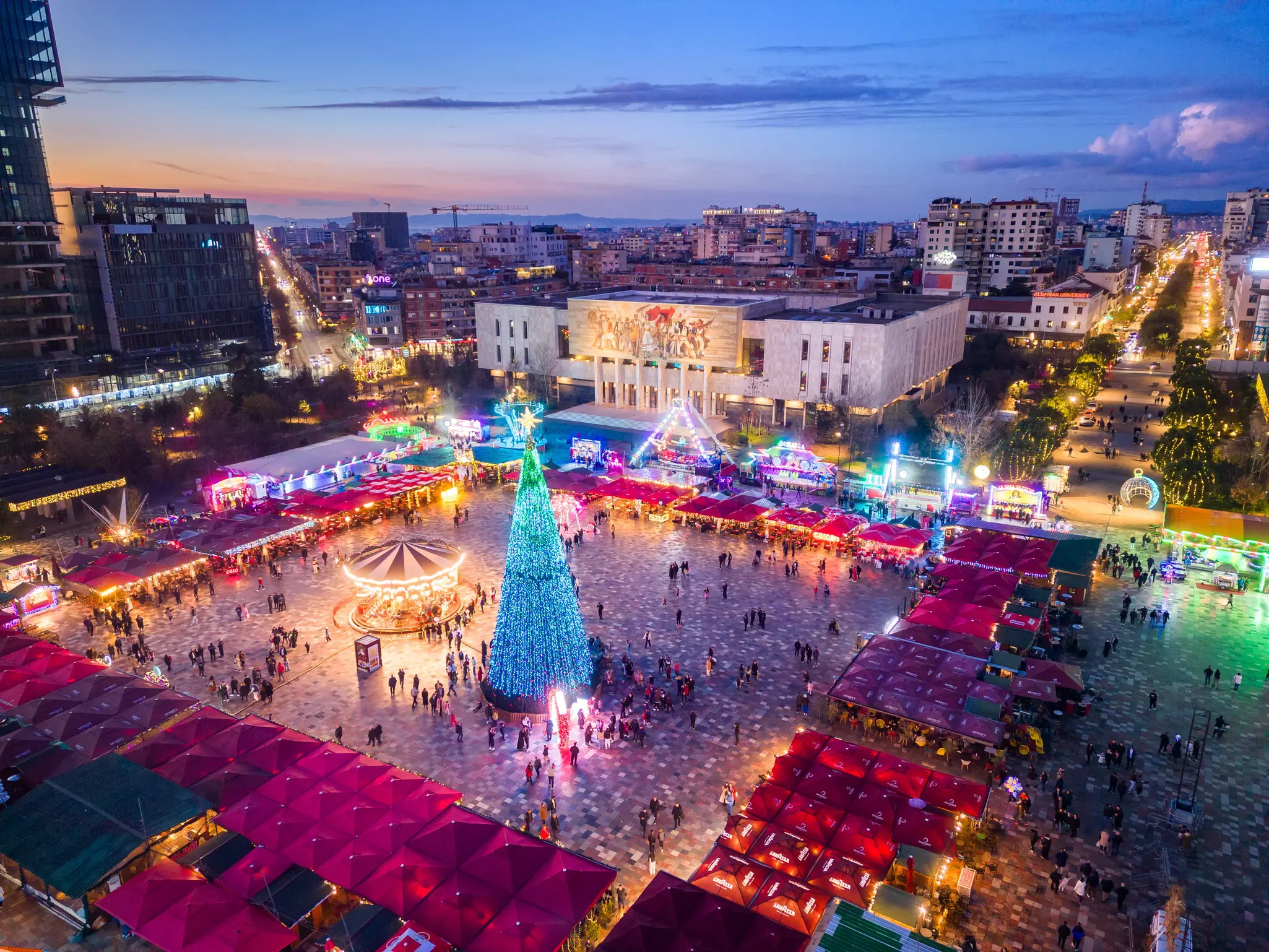 A city square lined with stalls and lit up in neon lights at dusk
