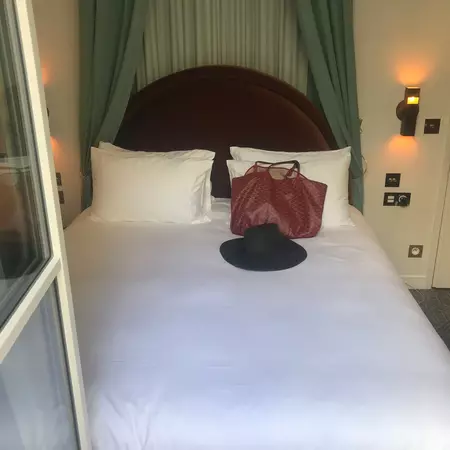 A bed with a white covering and pillows; a black hat and red bag are on the bed.