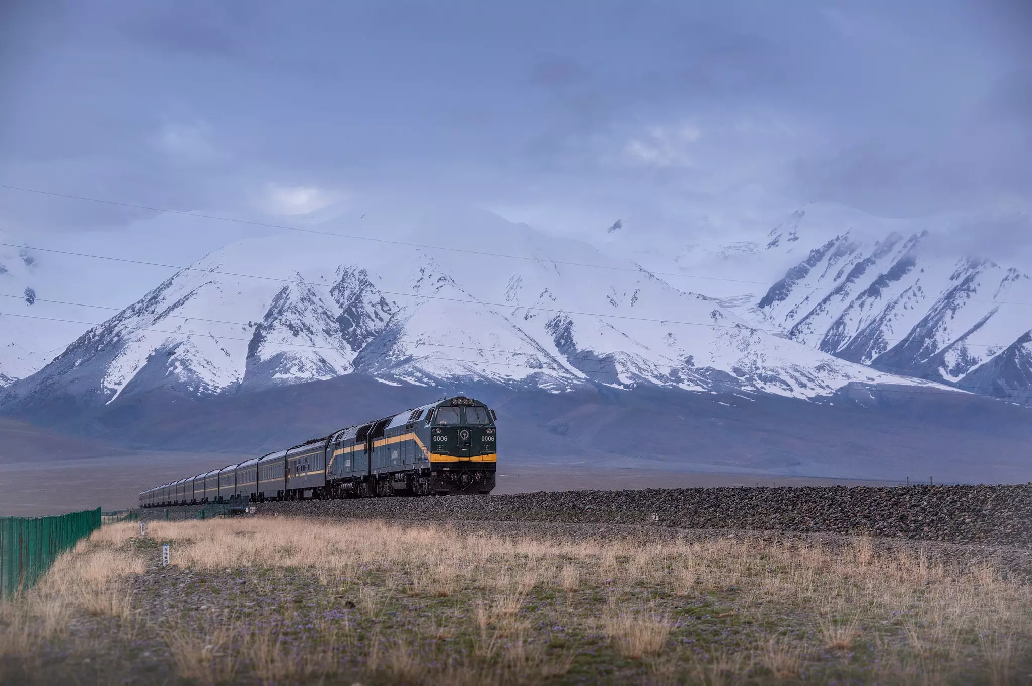 A train crosses a plateau, with dramatic, snow-covered mountains visible in the distance.
