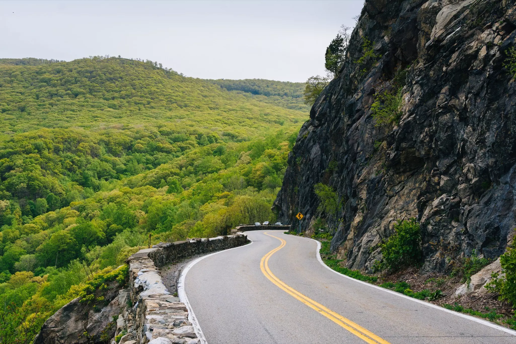 A two-lane highway in Cornwall on the Hudson, NY. On one side of the highway is a dense forested area. On the other side a tall mountain.