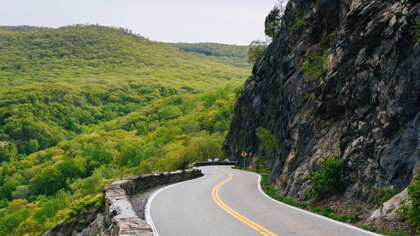 A two-lane highway in Cornwall on the Hudson, NY. On one side of the highway is a dense forested area. On the other side a tall mountain.