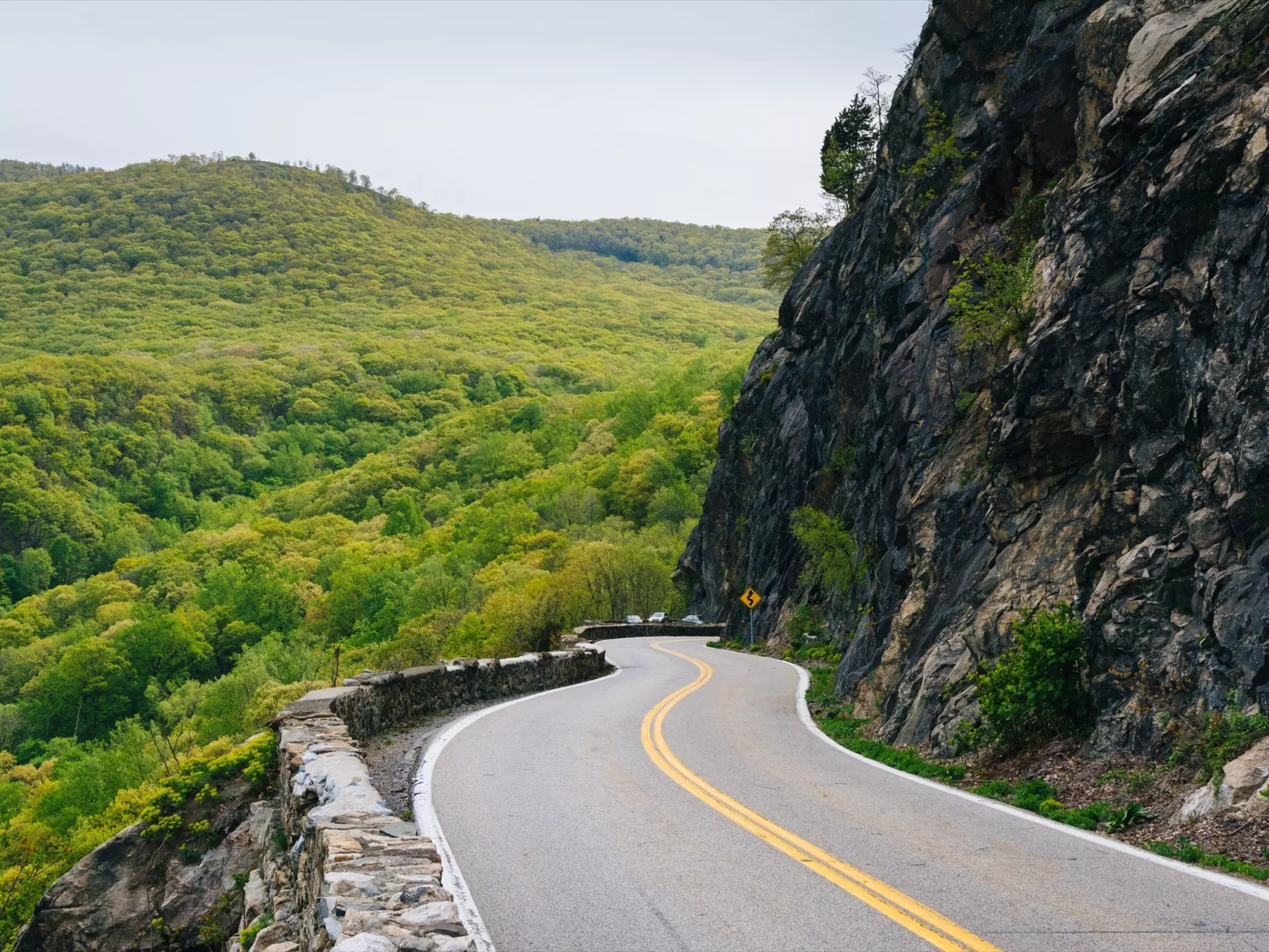 A two-lane highway in Cornwall on the Hudson, NY. On one side of the highway is a dense forested area. On the other side a tall mountain.