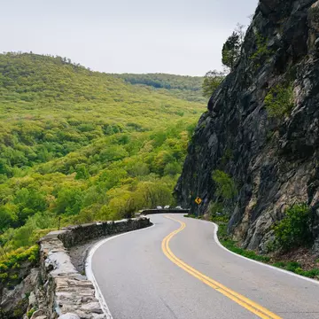 A two-lane highway in Cornwall on the Hudson, NY. On one side of the highway is a dense forested area. On the other side a tall mountain.