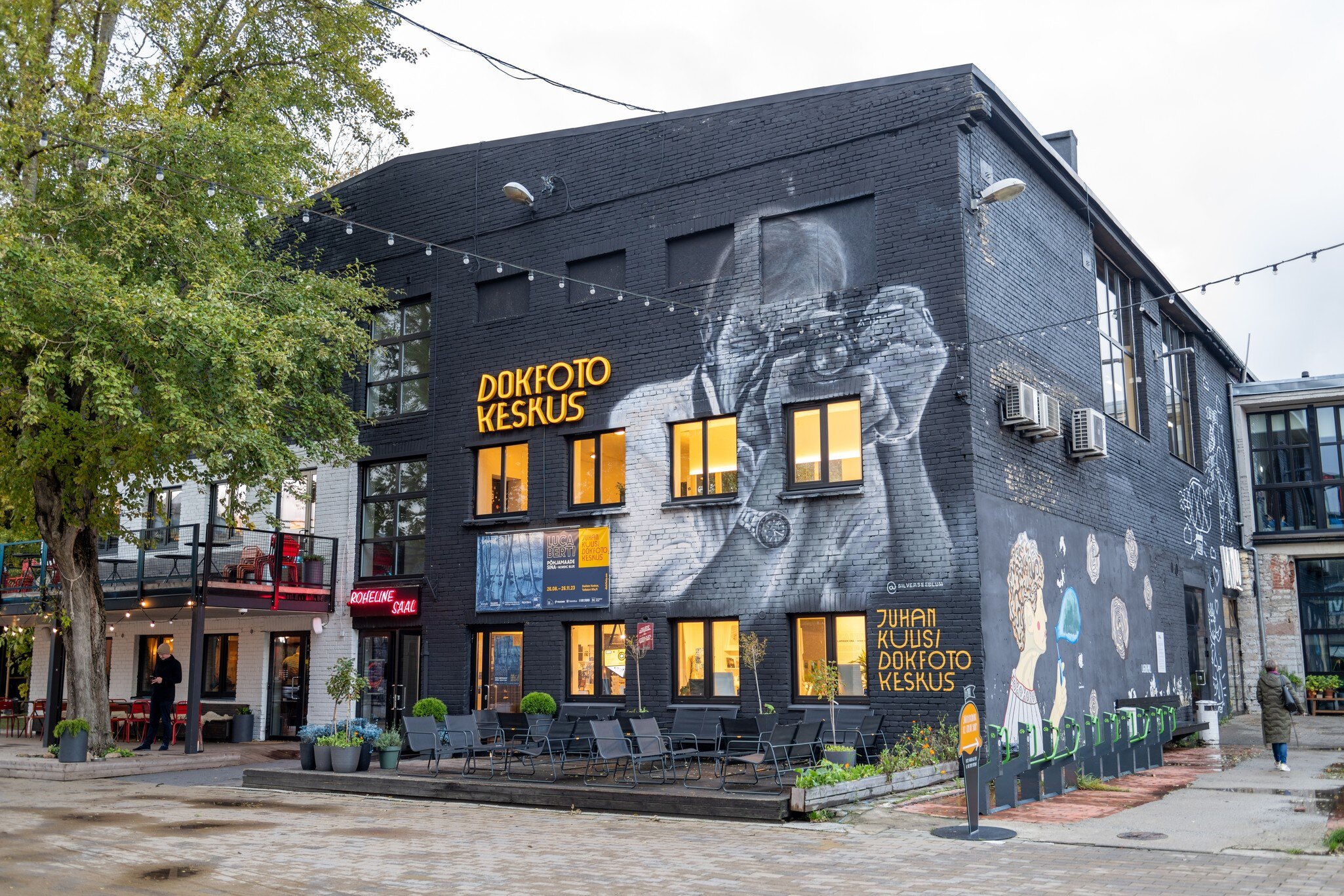 A dark building with a large black-and-white mural of a man with a camera, and outdoor cafe seating.