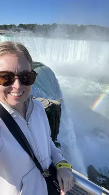 Woman in sunglasses and a tan sweater with the falls and a rainbow in the background. 