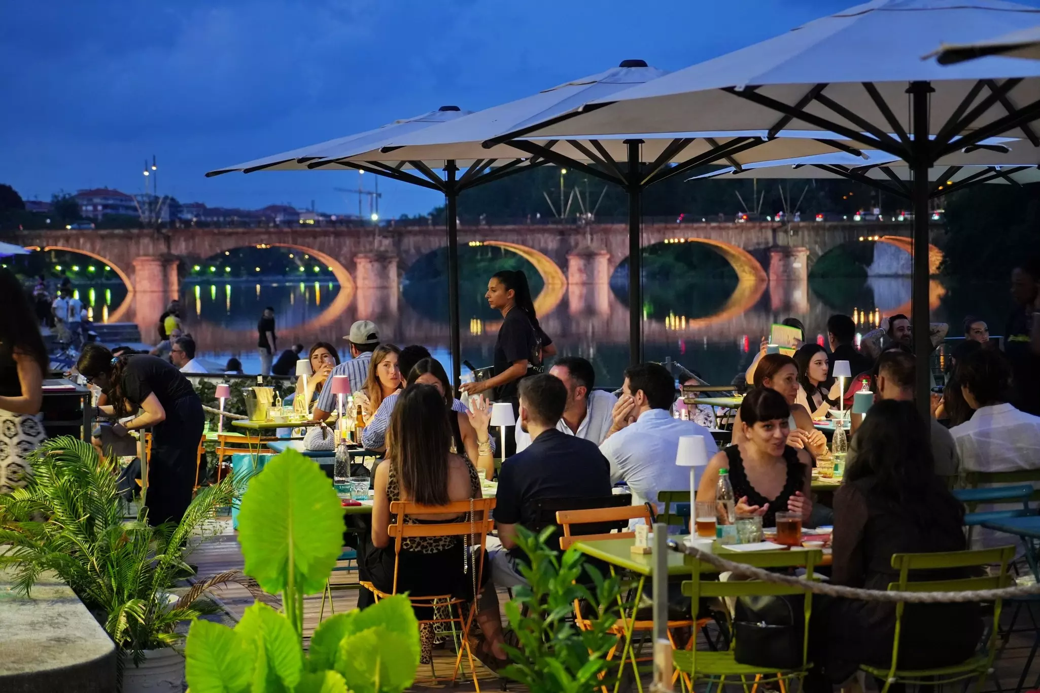 People sit at tables in an outdoor cafe by a river in the evening. The arches of a bridge in the background are underneath, and the reflections are cast in the water.