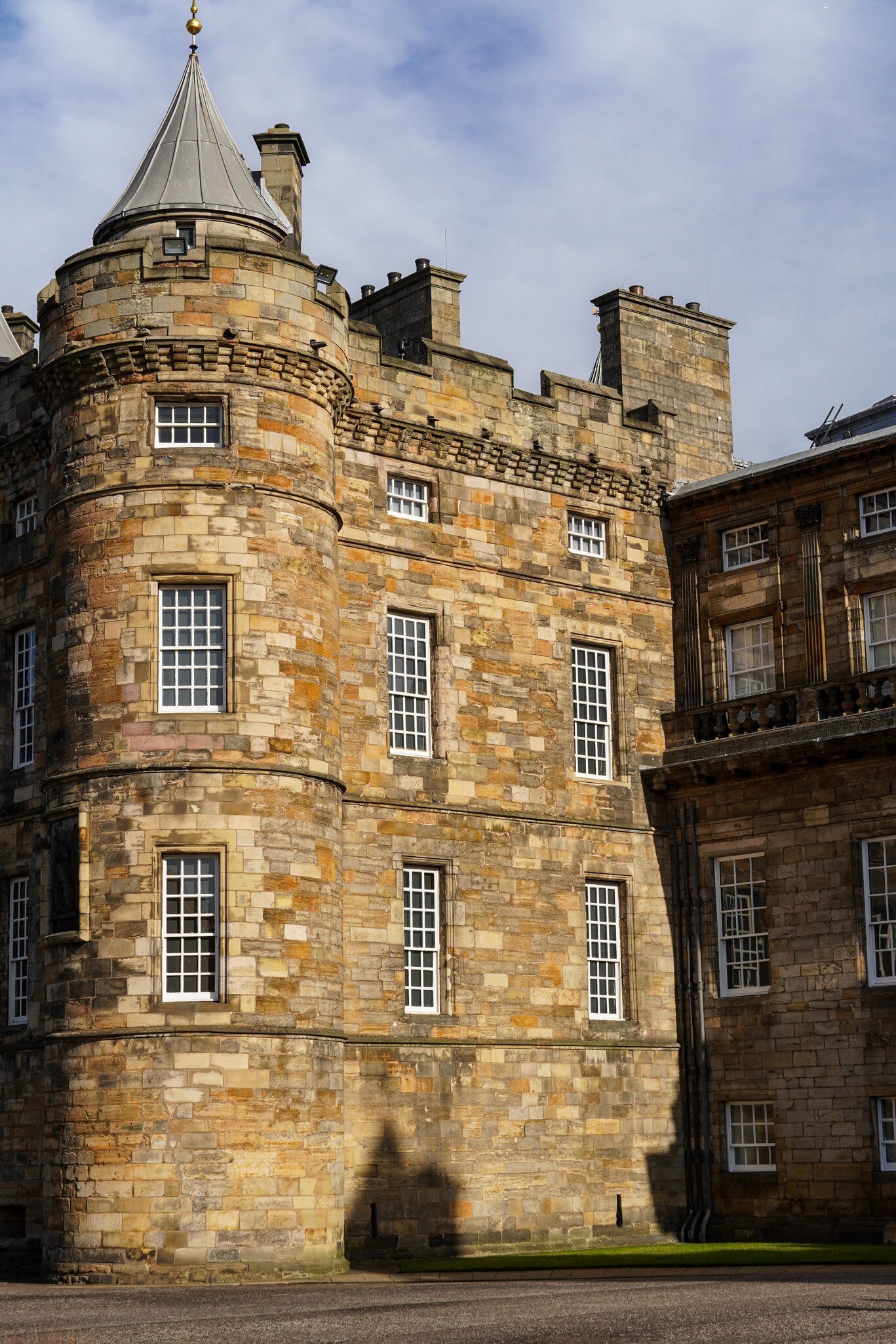 Holyroodhouse Palace Tower stands bathed in sunlight with dramatic shadows, highlighting its historic stone architecture and distinctive conical roof details.