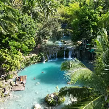Turquoise pool with a bamboo raft and swimmers beneath stepped waterfalls surrounded by palms, Siquijor, Philippines.
