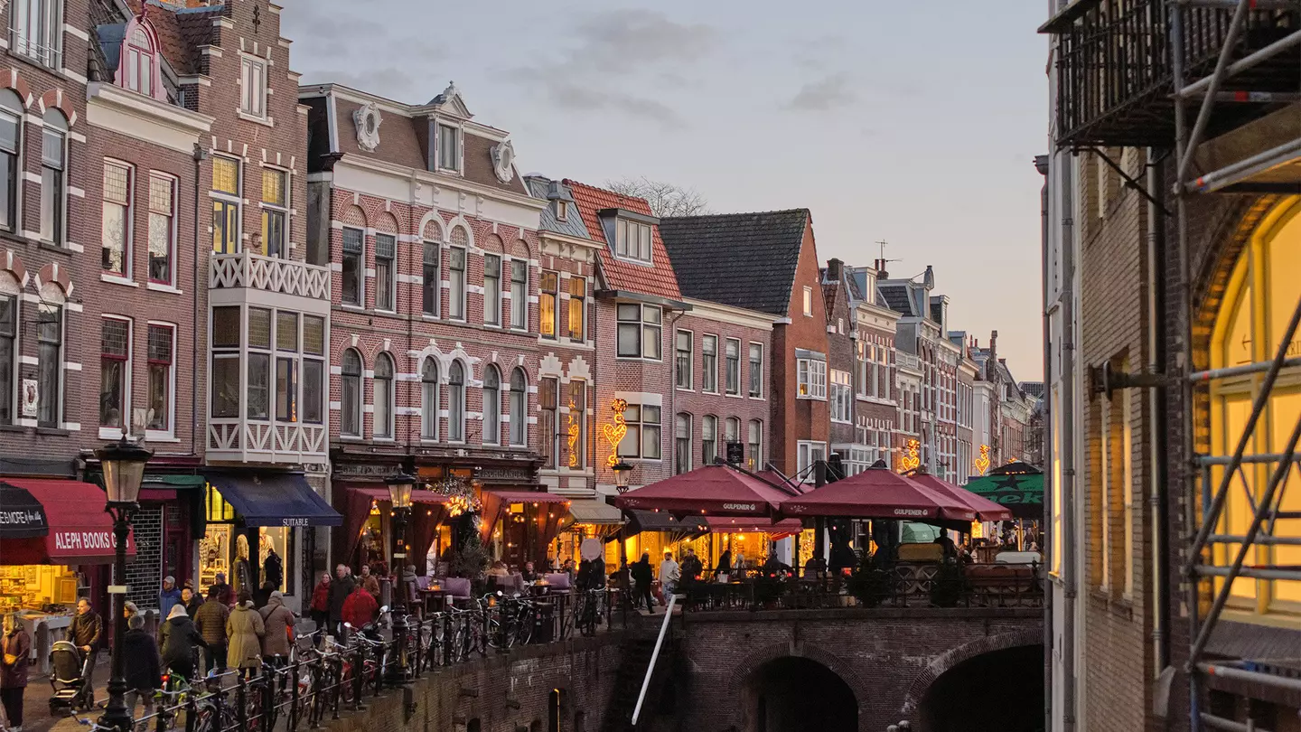 A canal lined with cafes, shops and bars; outdoor tables are under dark red umbrellas.