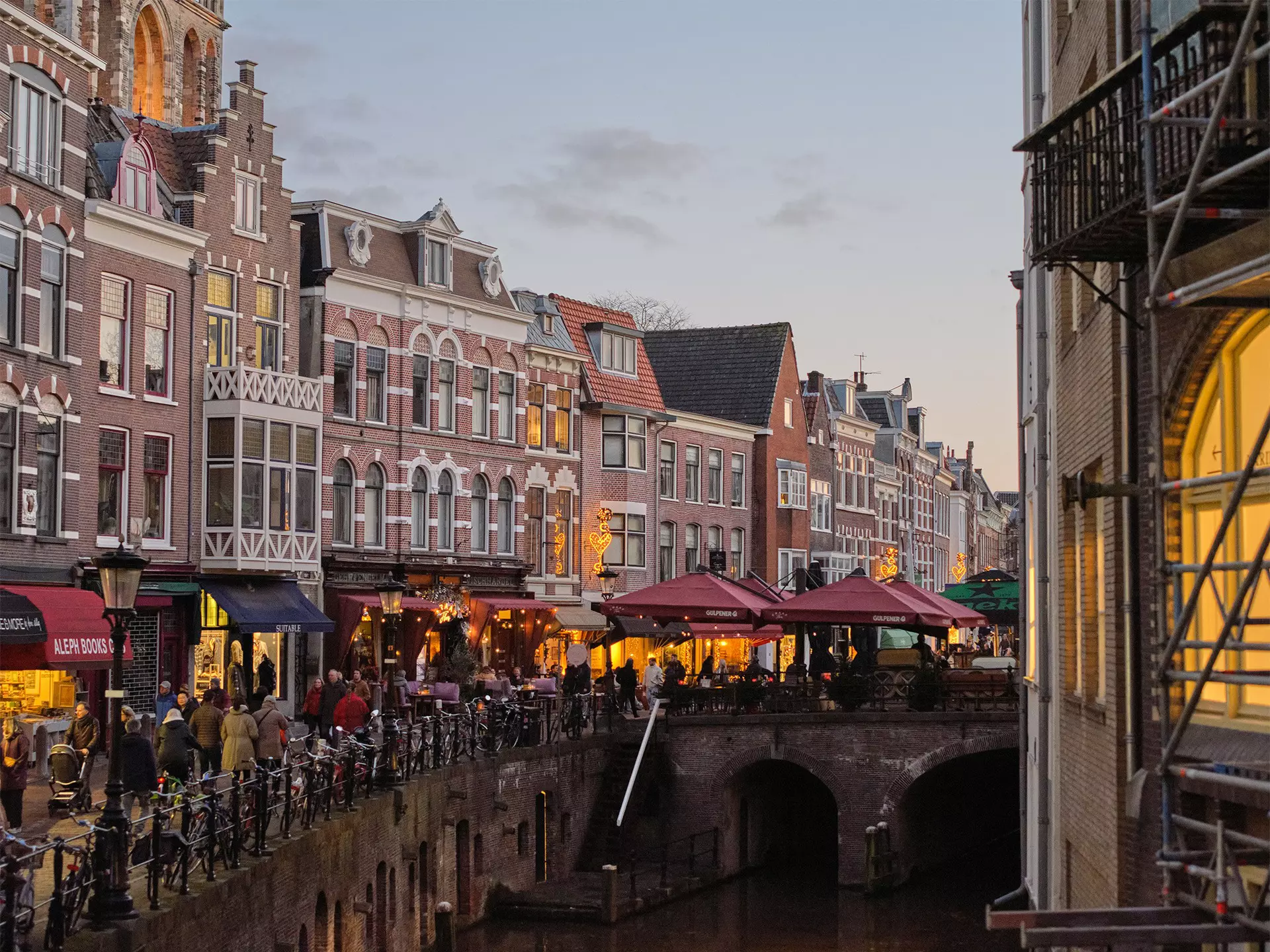 A canal lined with cafes, shops and bars; outdoor tables are under dark red umbrellas.