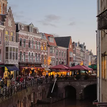 A canal lined with cafes, shops and bars; outdoor tables are under dark red umbrellas.