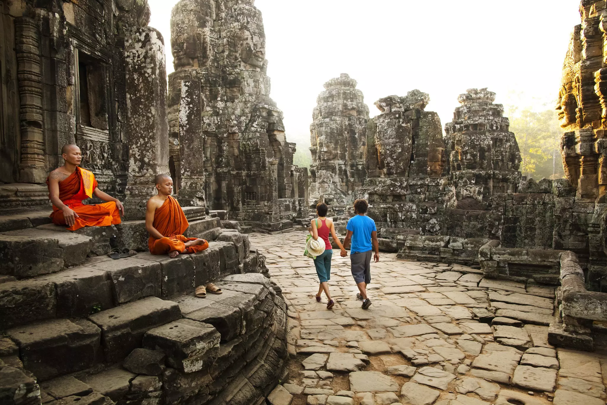 Two visitors walk through a temple complex, past two monks in saffron robes who are seated in prayer.