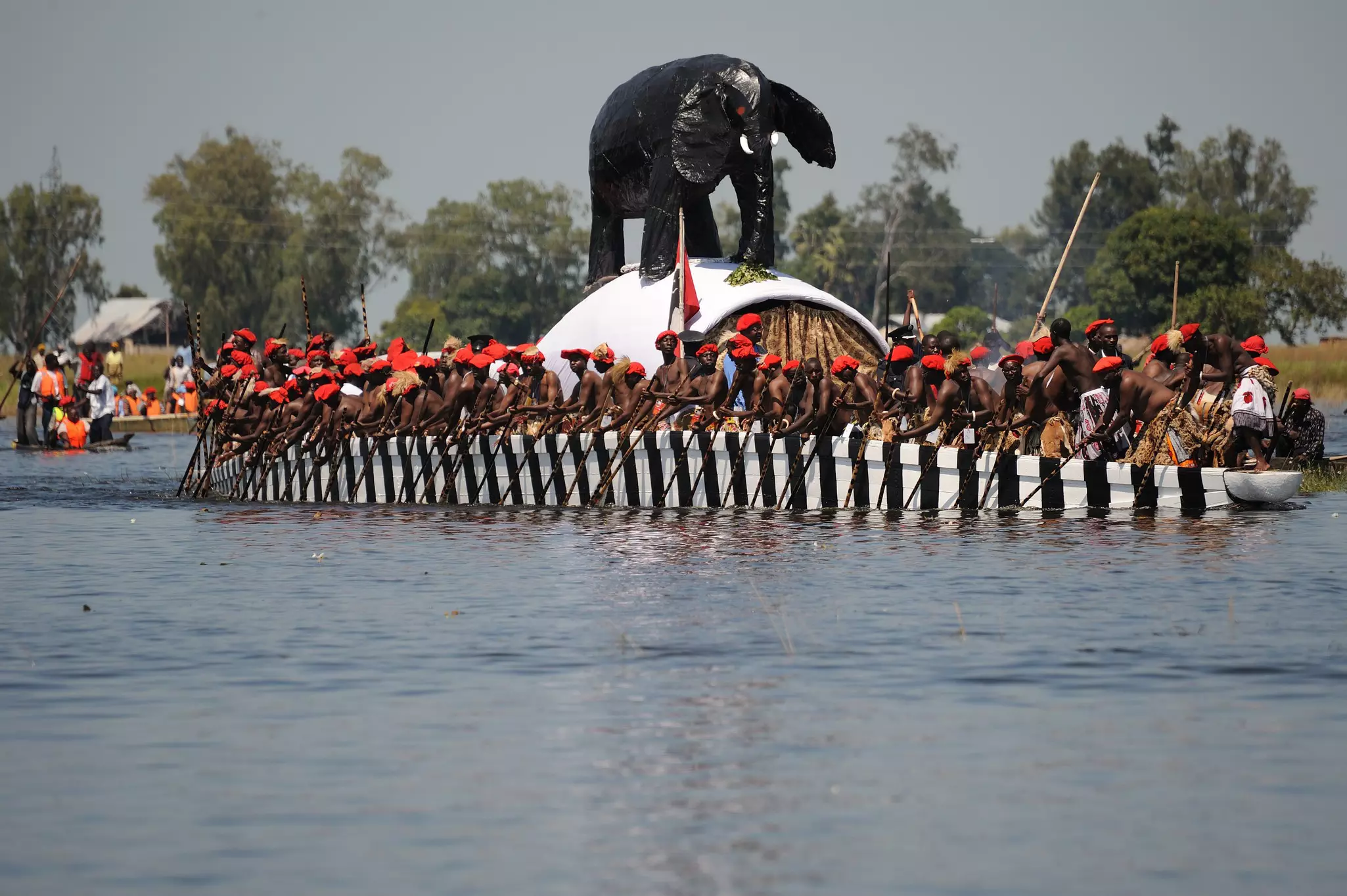Group of men in red berets rowing a long, black and white striped boat with a black elephant figure on top of it across a body of water