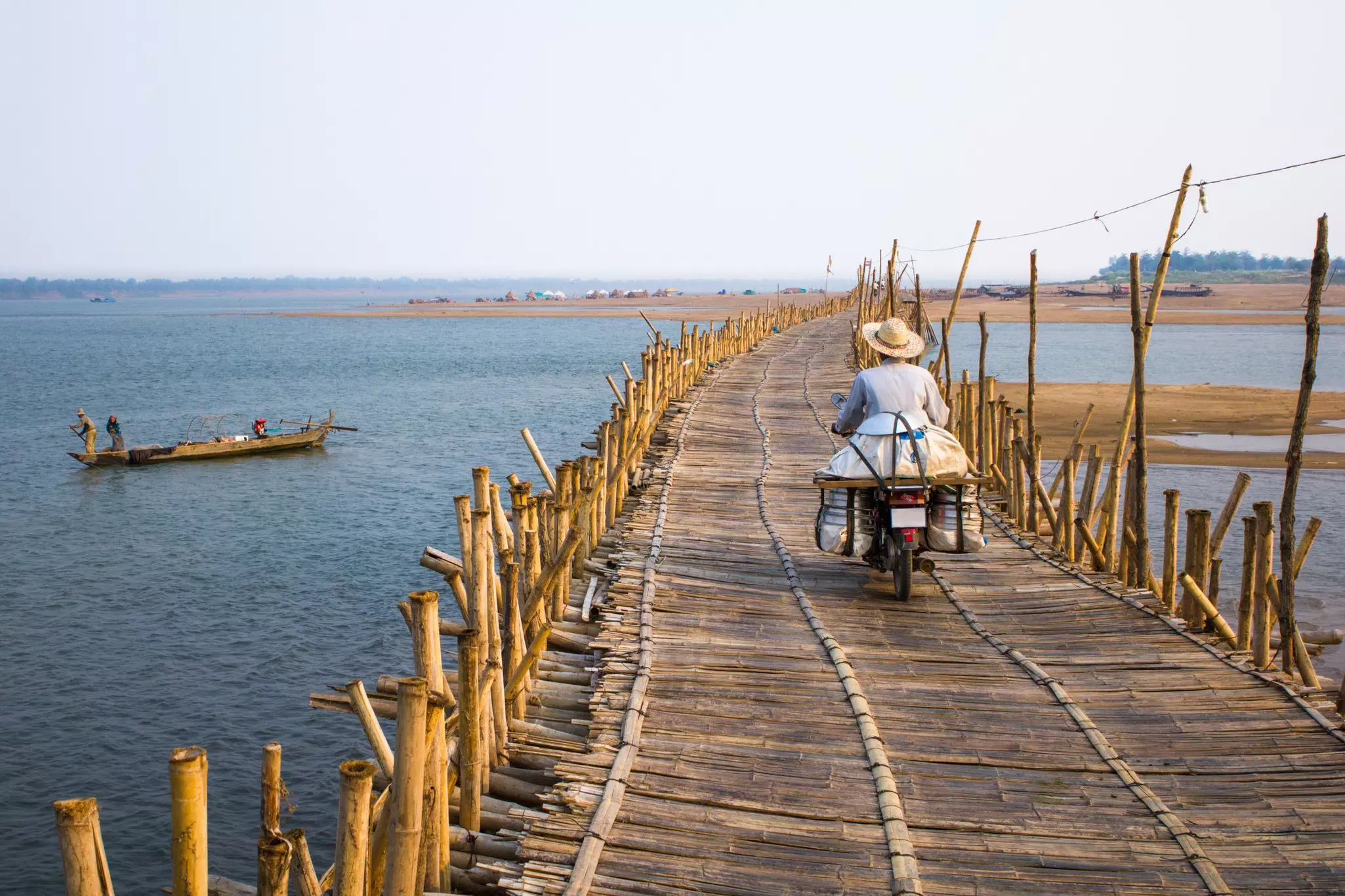 A person on a motorbike crossing a bamboo bridge over a river in Cambodia; there is a small boat with fishers to the left.