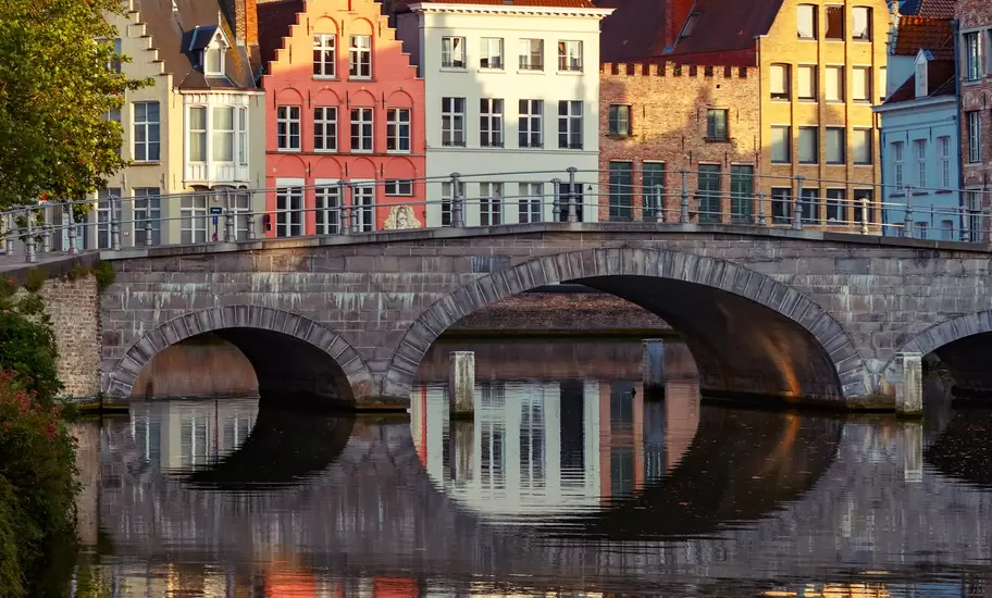 Bruges canal with a stone arched bridge and stepped-gable houses reflected in still water, a church tower above