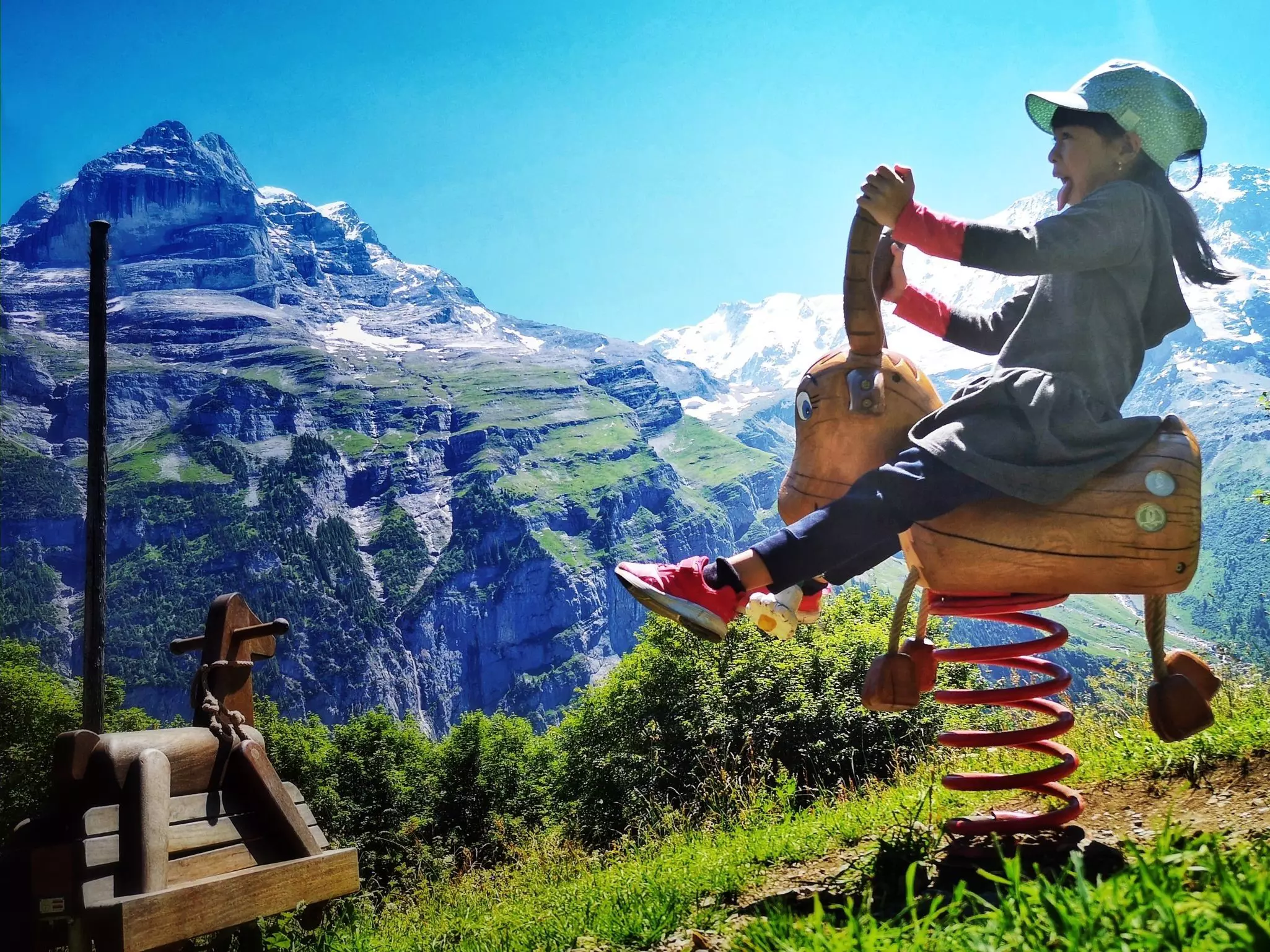A little girl with long black hair under a bright green hat sticks her tongue out with excitement as she rides a wooden horse playground toy in view of a large, snow-dusted mountain.
