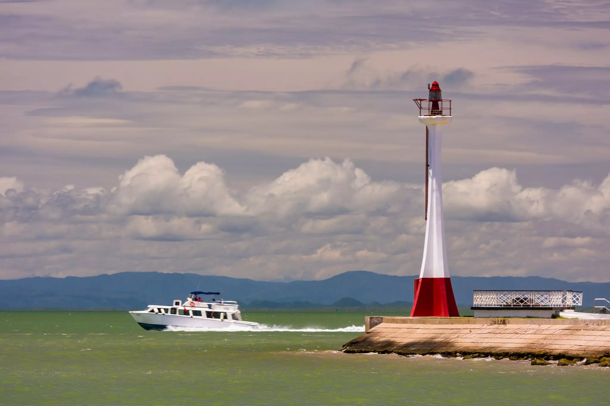 A power boat moves through greenish water past a narrow red-and-white lighthouse on a cloudy day.