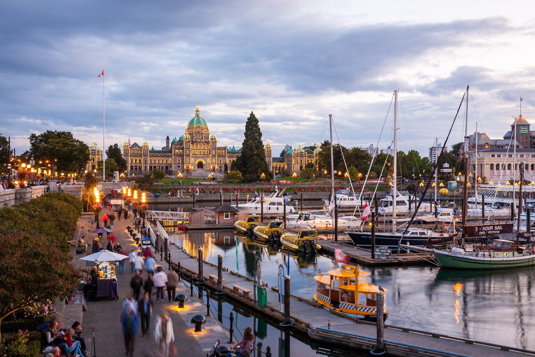 Illuminated domed parliament building at dusk overlooking an inner harbour marina lined with sailboats and strolling crowds