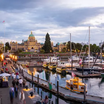 Illuminated domed parliament building at dusk overlooking an inner harbour marina lined with sailboats and strolling crowds