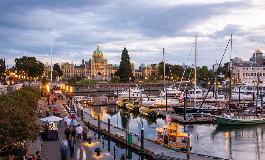 Illuminated domed parliament building at dusk overlooking an inner harbour marina lined with sailboats and strolling crowds