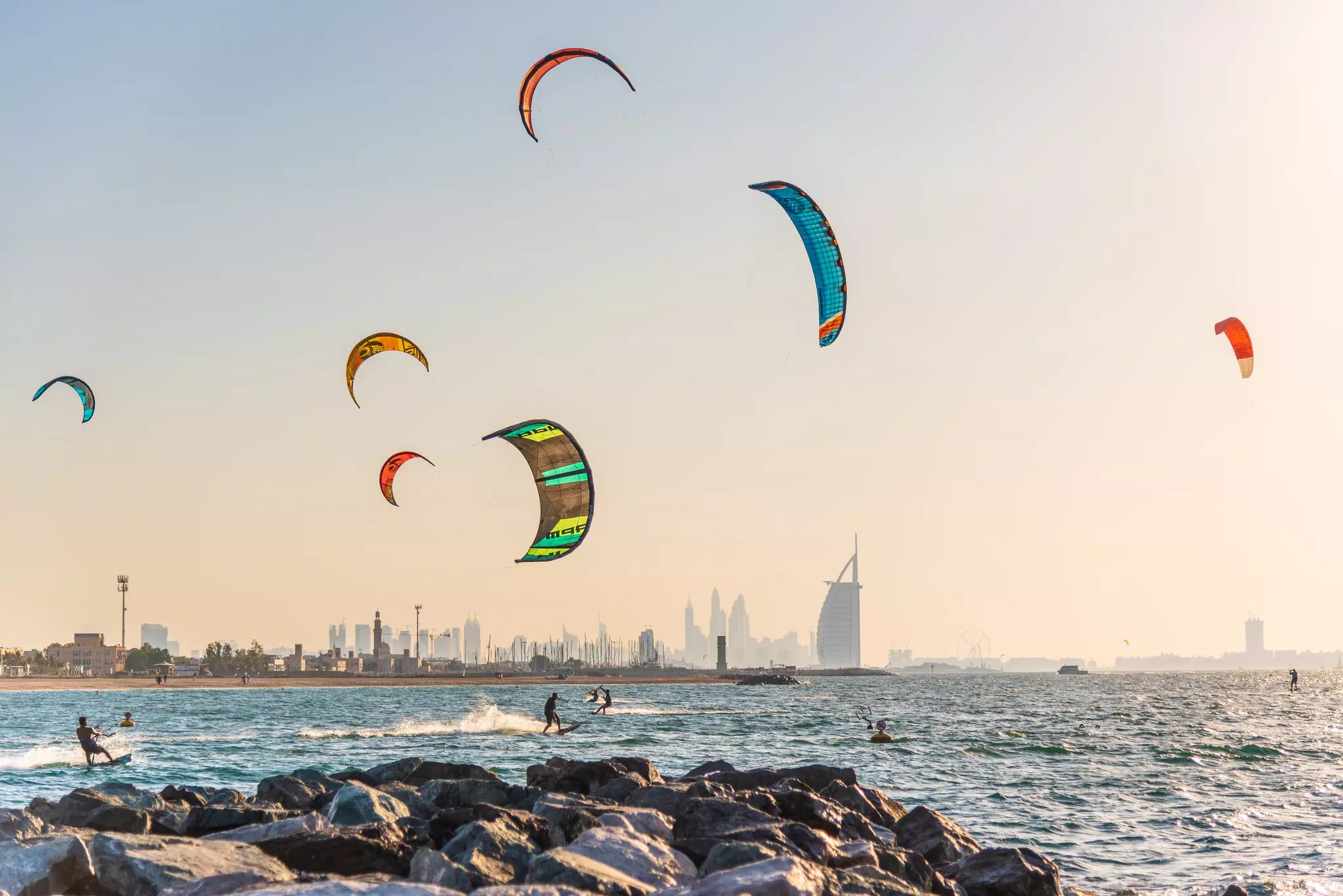 The sails of kitesurfers fill the sky off a beach in a city. The silhouettes of skyscrapers are seen in the distance.