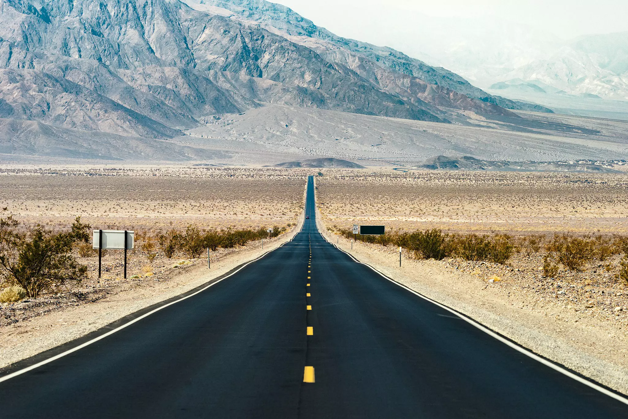 A road of smooth black asphalt disappears in to the distance in a desert landscape heading towards hills.