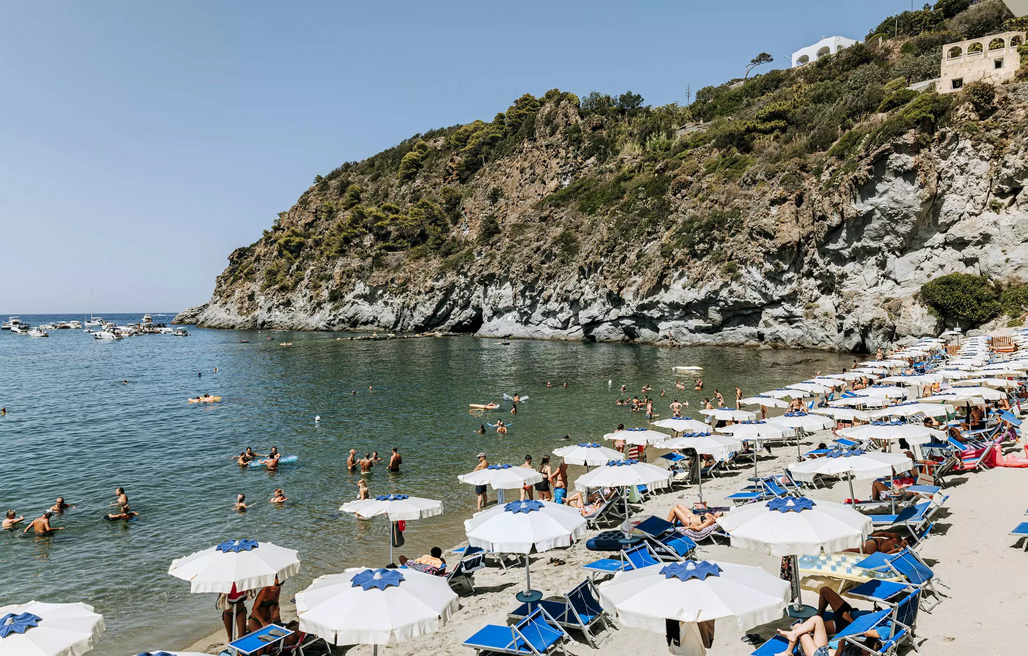 A beach is filled with sun loungers and umbrellas, with people swimming just offshore. A steep slope at the edge of the beach leads to the water.