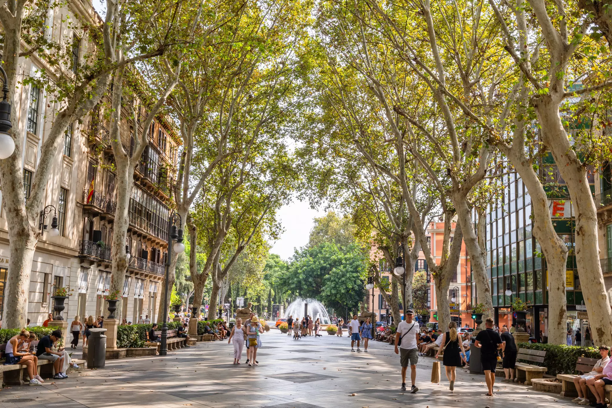 People walk on a shaded promenade with a fountain at the end.