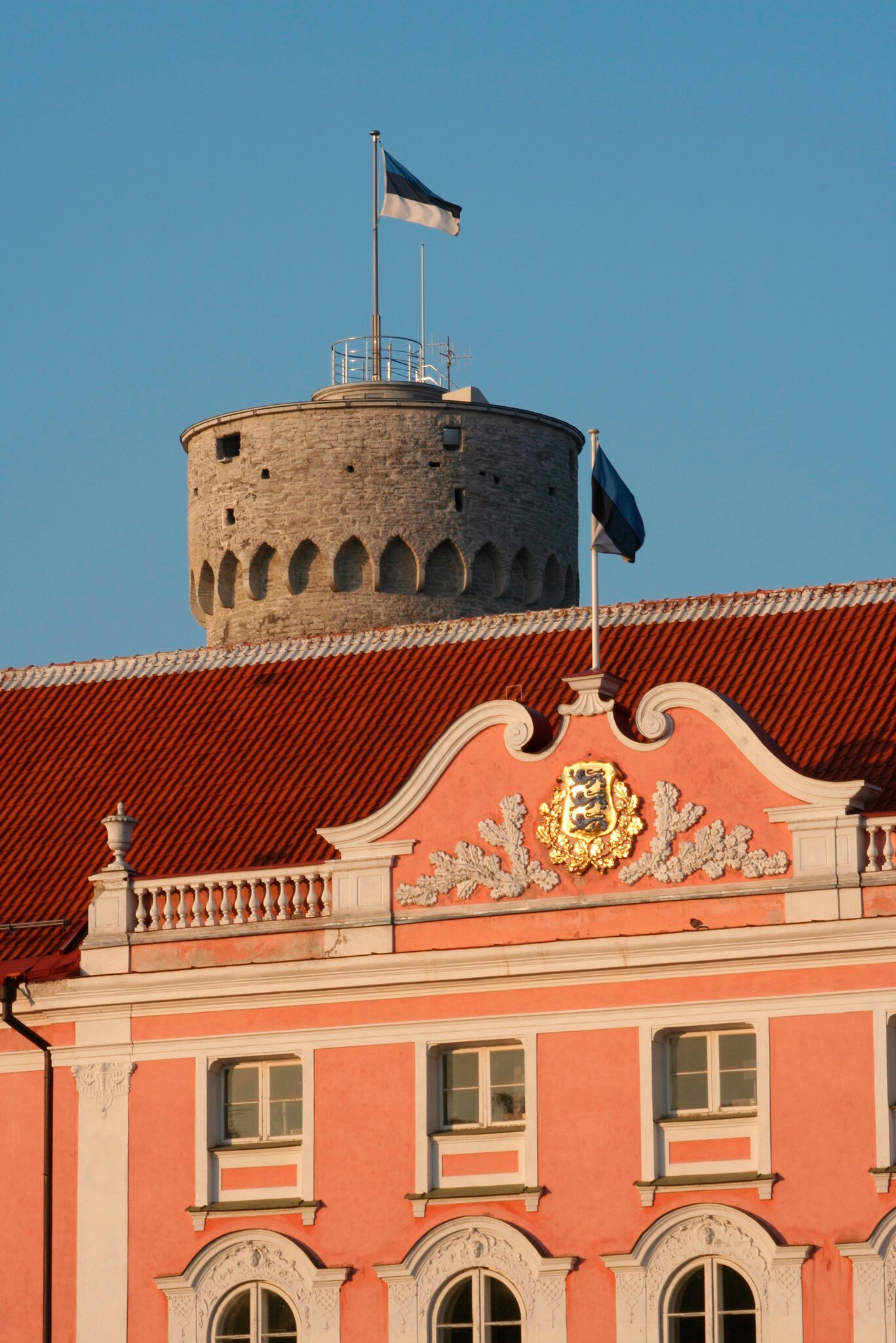 Pink baroque palace facade with a gilded coat of arms, a round stone tower and national flag rising behind it.