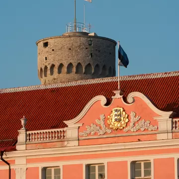 Pink baroque palace facade with a gilded coat of arms, a round stone tower and national flag rising behind it.
