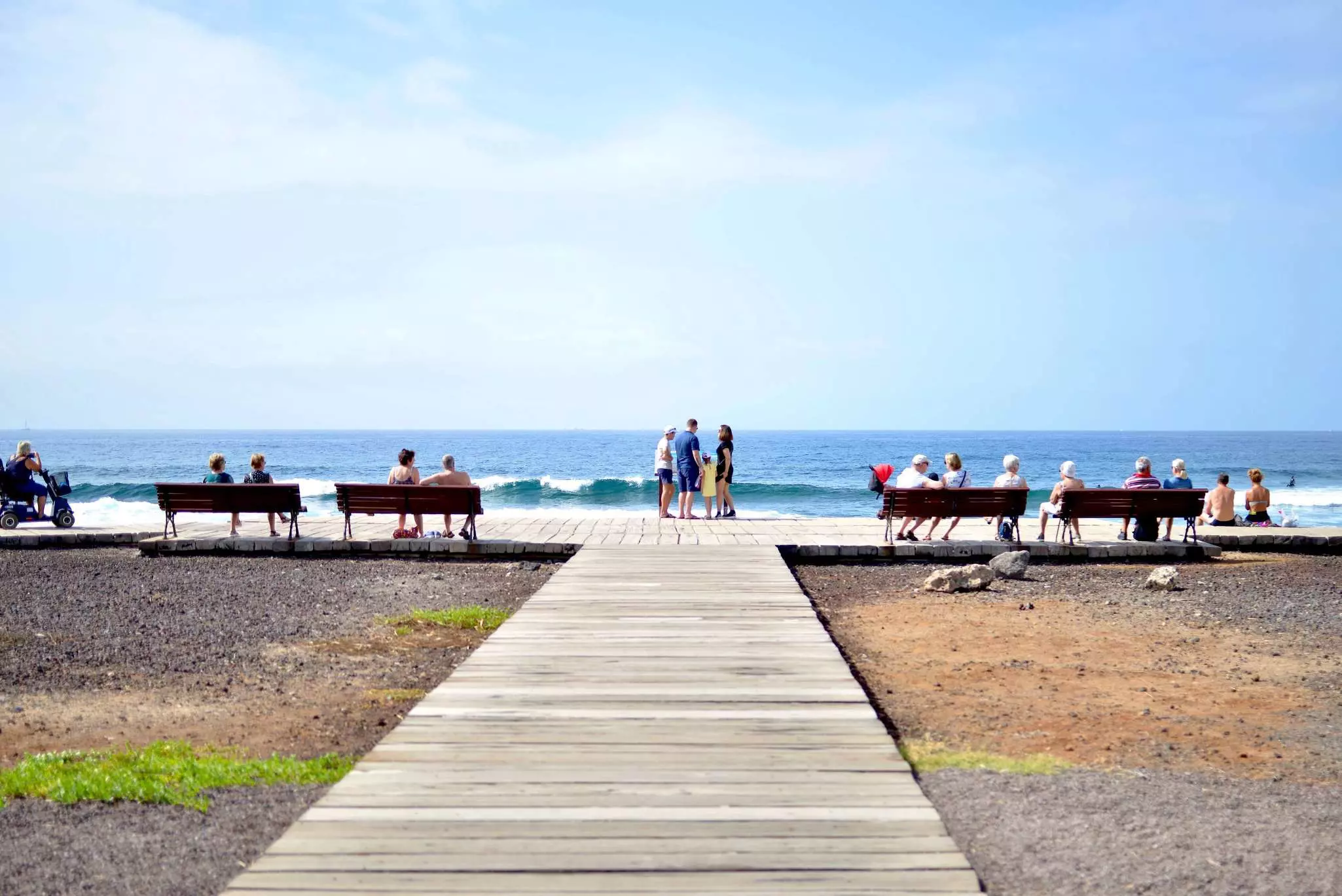 People relaxing on benches on a promenade in front of waves on a beach with blue sky and sea
