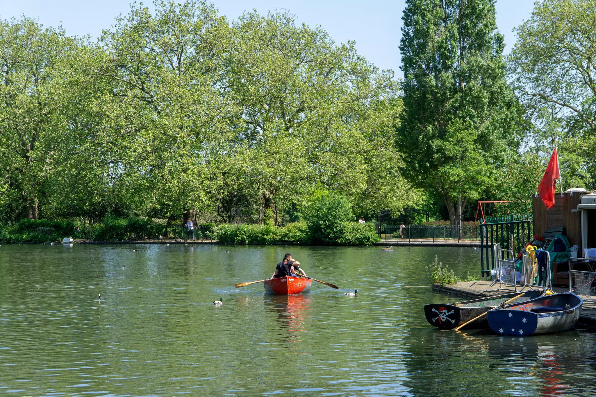 A man rows a red boat through a lake surrounded by a concrete walking path and trees, with two boats docked to the right outside a low building on a sunny day.