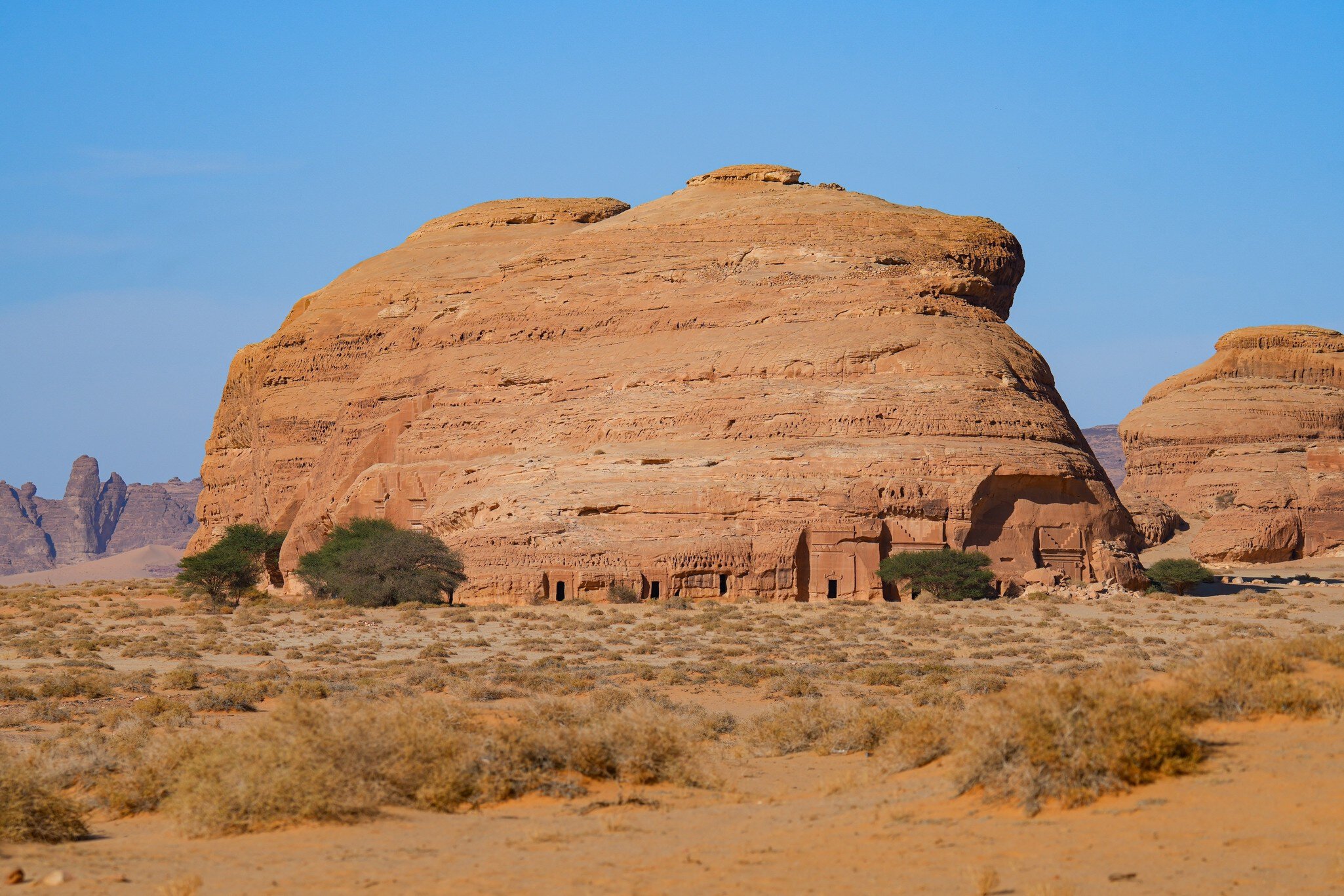 A reddish rock in an open desert plain with tombs carved into the base.