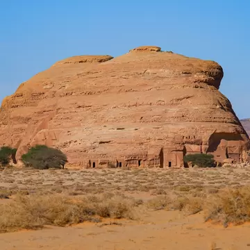 A reddish rock in an open desert plain with tombs carved into the base.
