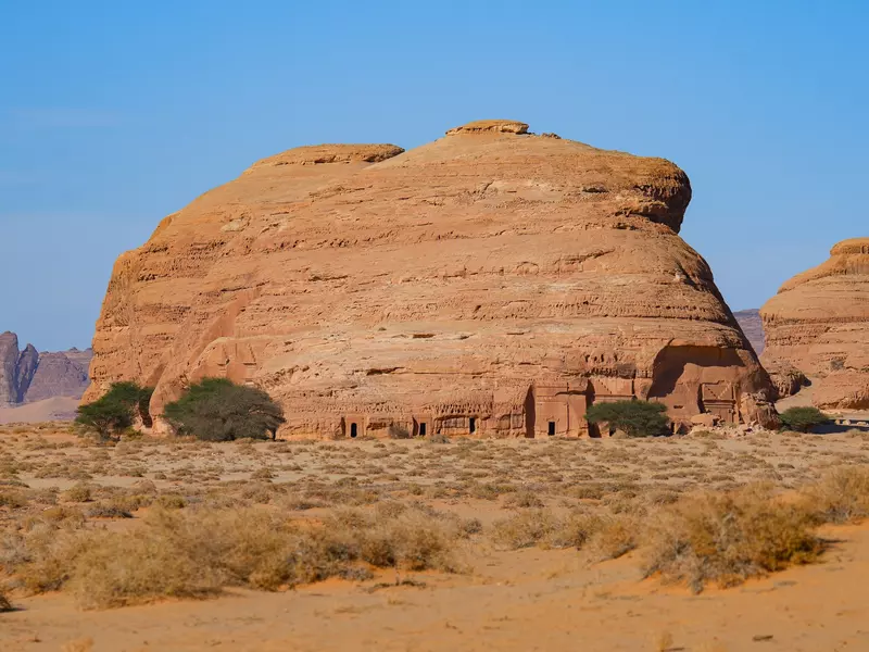 A large rock in a desert landscape has doorways to tombs at its base.